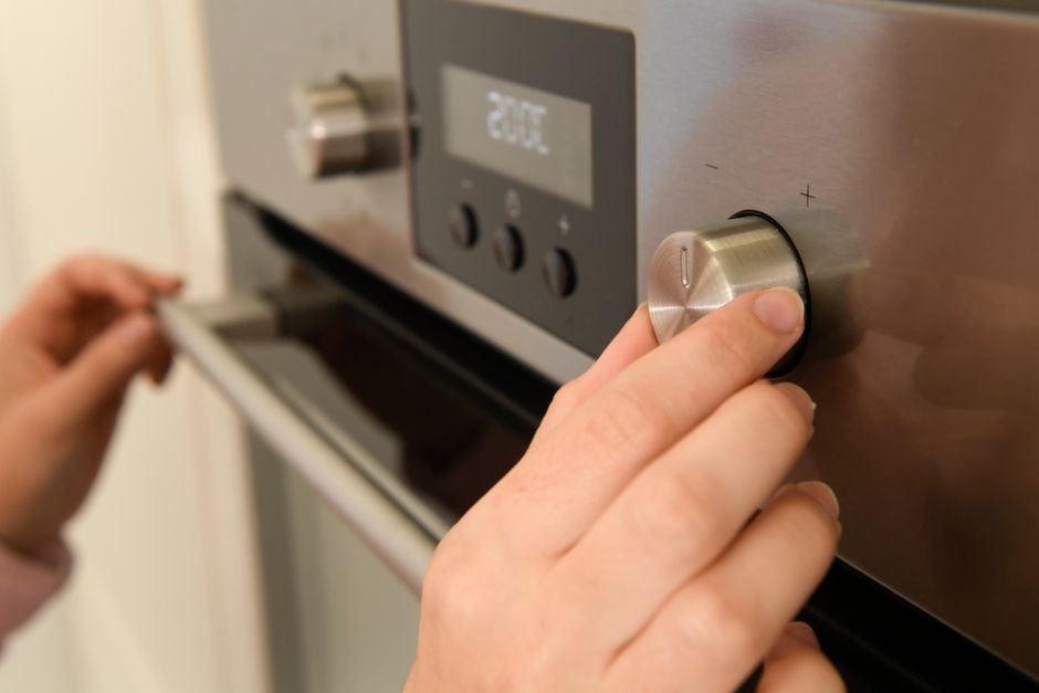 A woman wearing a white t-shirt, yellow and blue cleaning gloves, is leaning forward and inspecting the interior of a black oven with a glass door in a modern kitchen. She holds a spray bottle in one 