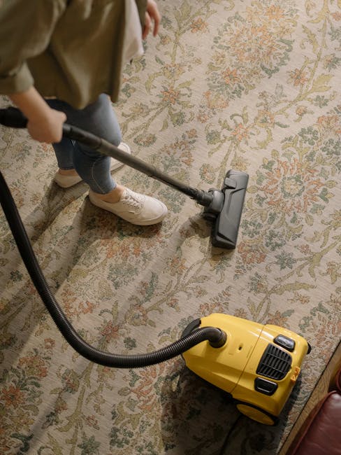A woman is engaged in surface cleaning of a living room area, using a yellow vacuum cleaner with a hose attachment to deep clean a patterned, beige and blue rug. She is dressed casually in a beige jac